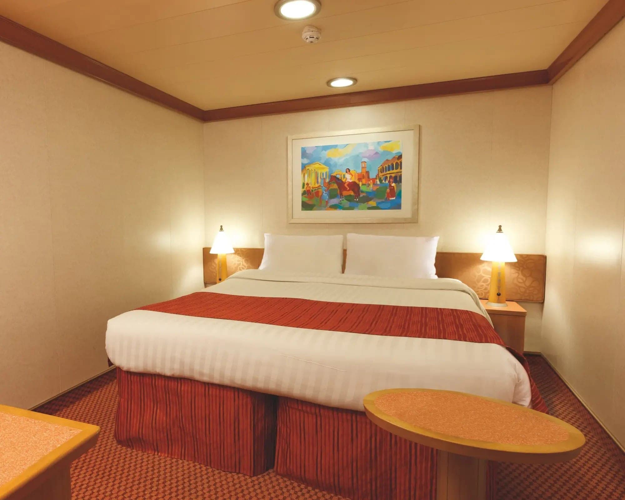 Cabina interna del Costa Fascinosa: cama matrimonial con cubrecama rojo, dos veladores y un cuadro, sin ventana