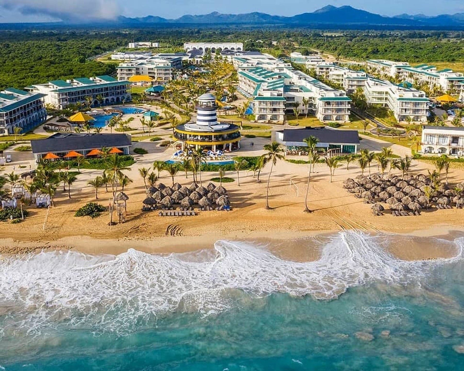 Vista aérea del hotel Ocean El Faro en Punta Cana: los edificios, las piletas, el faro y la playa con sombrillas de paja frente al mar