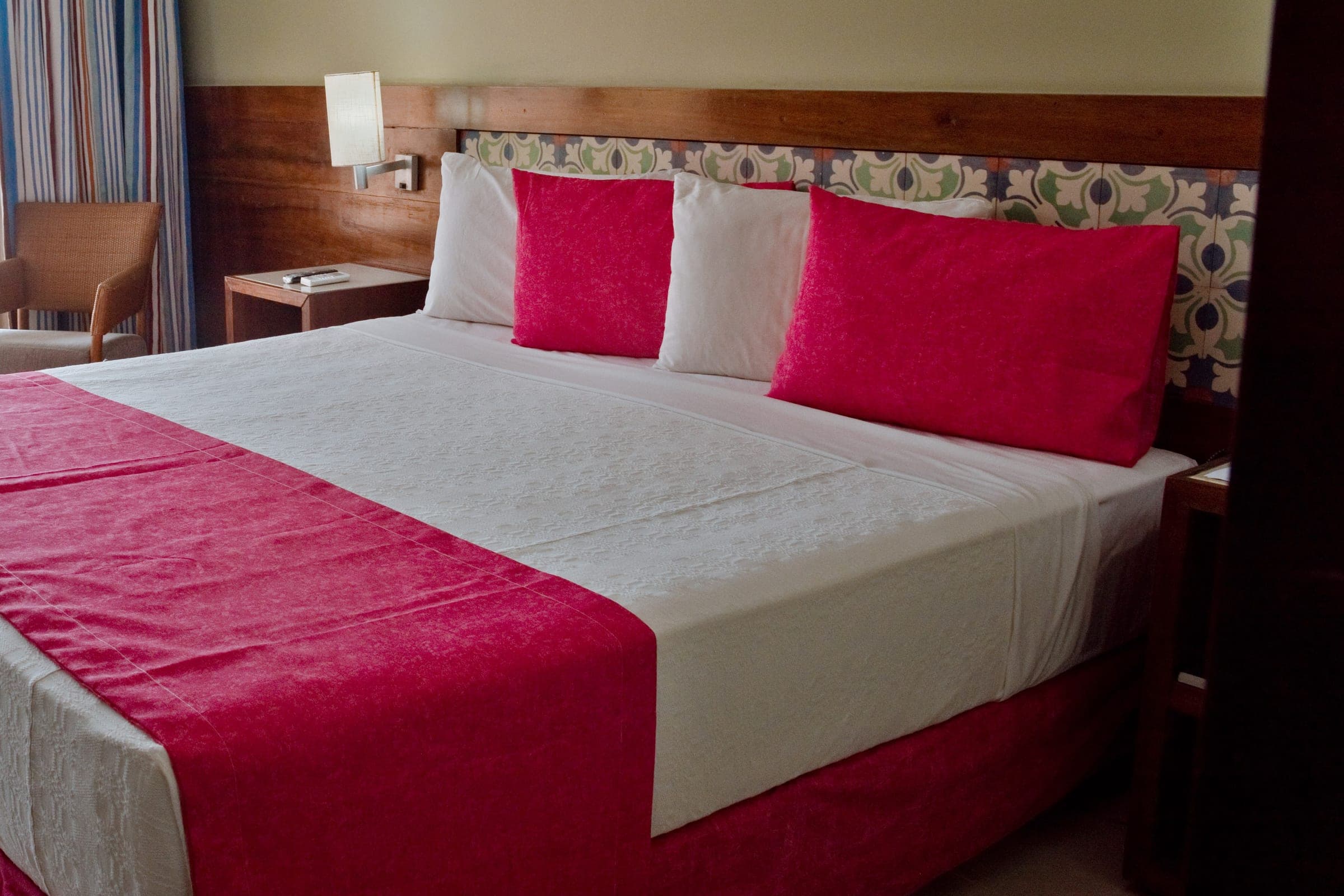 Habitación del hotel con cama matrimonial de almohadones rojos, respaldo de madera y azulejos, y una mesa de luz