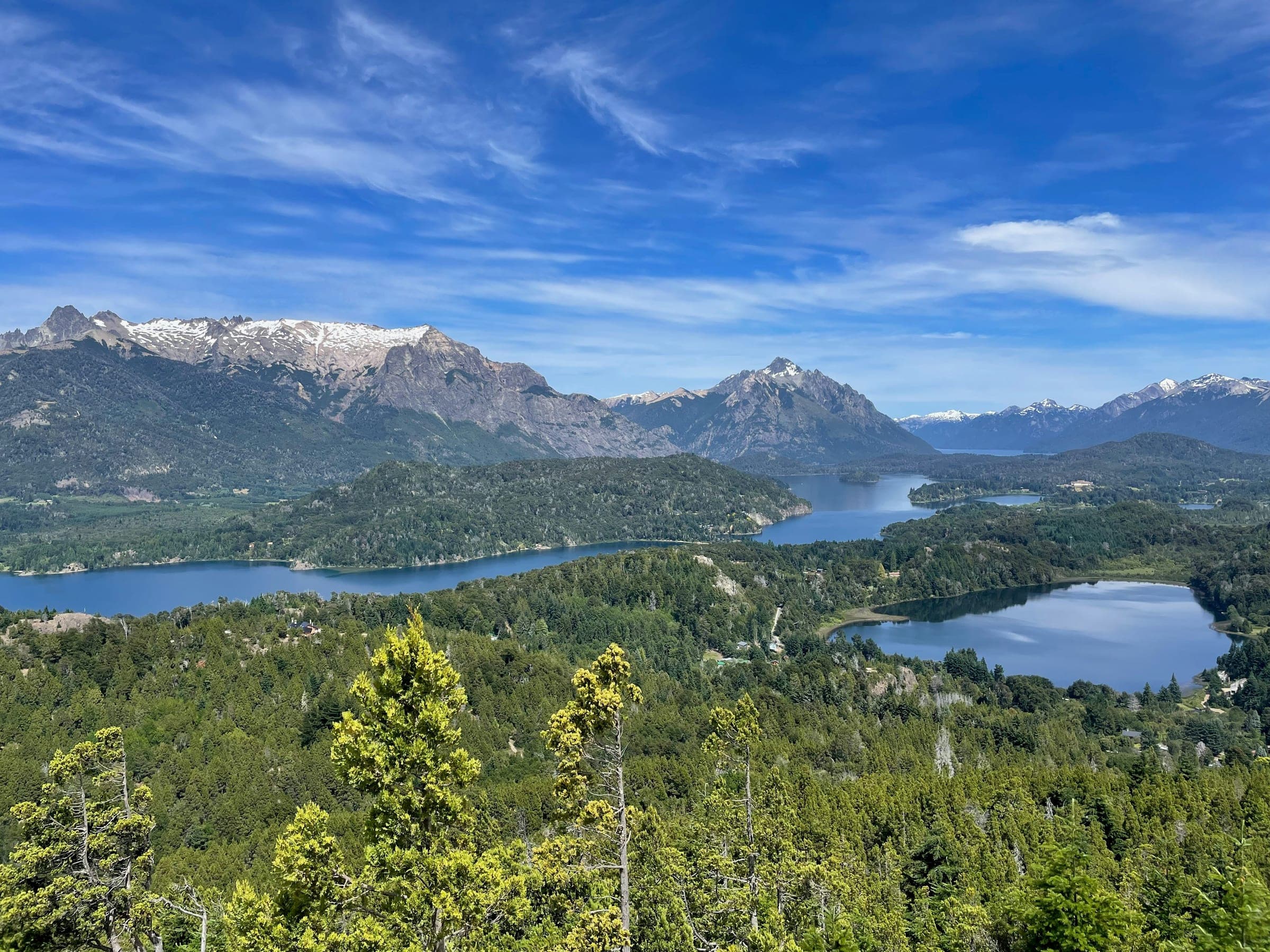 Dos lagos azules entre penínsulas de bosque y un cordón de montañas nevadas, vistos desde un cerro en Bariloche